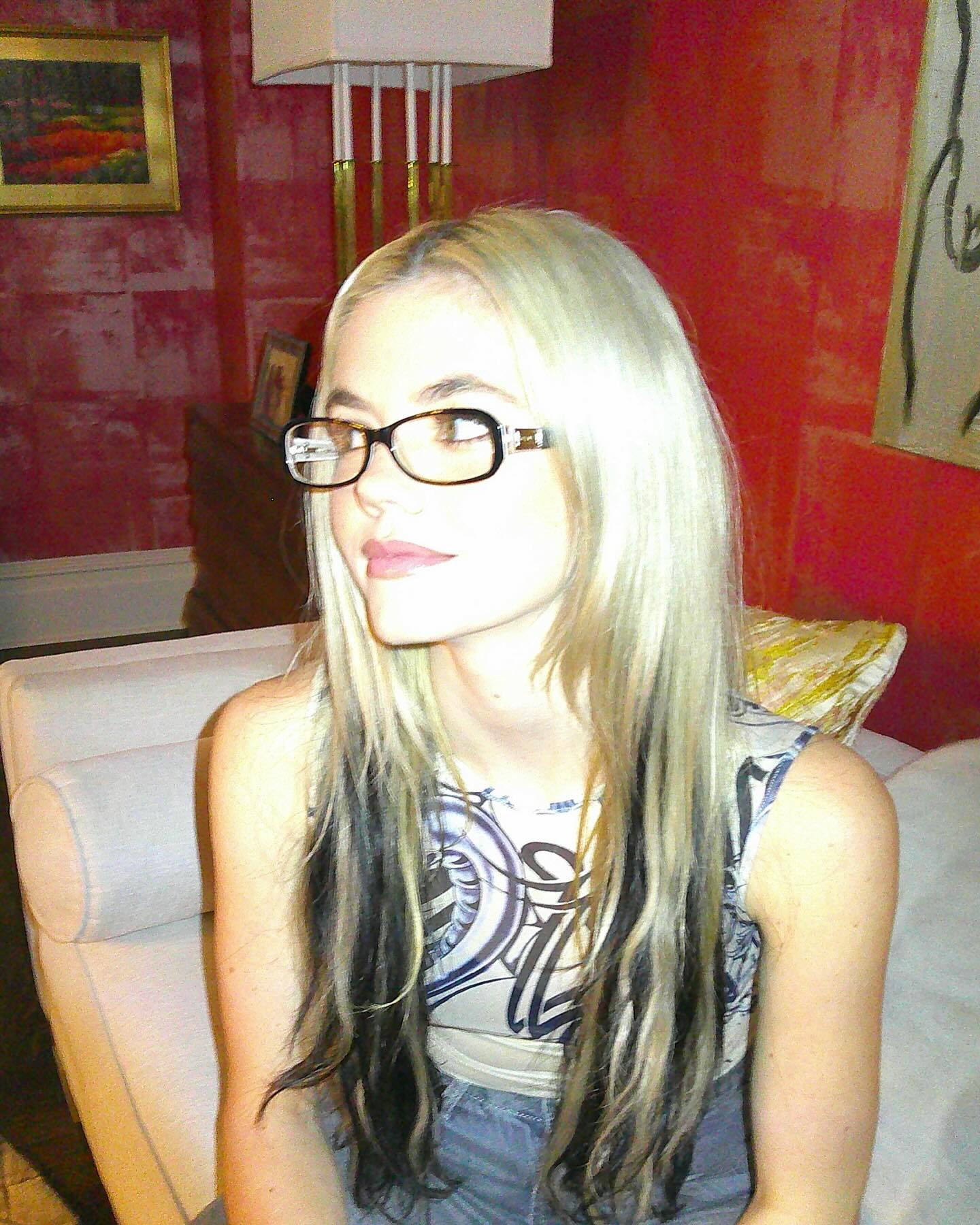 a woman with blonde and black hair is sitting indoors wearing eyeglasses and a tank top. there is a painting on the wall behind her, a lamp, and other furniture in the room.