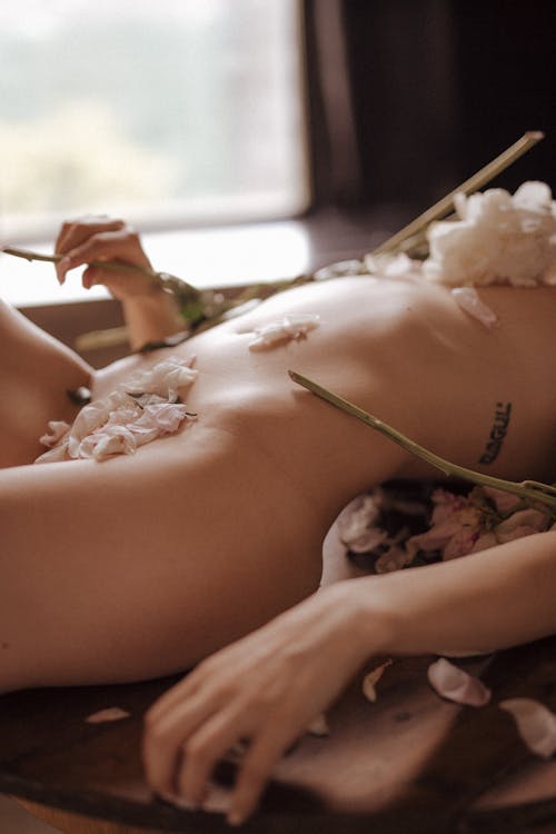 the image is a closeup shot of a nude woman lying on her side.  her body is adorned with delicate, light pink and white flower petals and stems, predominantly concentrated around her lower abdomen and pubic area. the petals are strewn across her body, creating a visually striking contrast against her pale skin. her hands are relaxed, one gently holding a stem, the other resting on a surface beside her. soft, natural light from a window illuminates the scene, creating a warm and intimate atmosphere.  a small tattoo is partially visible on her rib cage. the overall mood is serene and sensual. the focus is soft, lending a dreamlike quality to the photograph.
