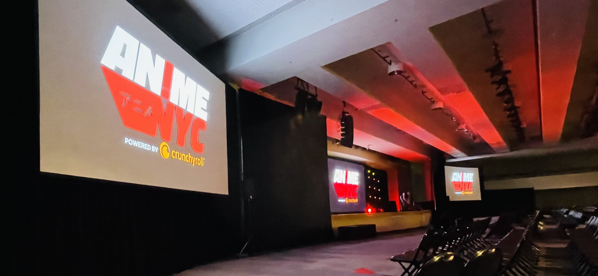this is a wide, indoor shot of a mostly empty auditorium set up for anime nyc. there is a stage in the background with three screens displaying the anime nyc logo, which is also projected onto a larger screen on the left. the primary lighting is red and focused on the stage, with the rest of the seating area dimly lit. there are rows of black folding chairs on the right, and a few people can be seen on the stage in the distance.