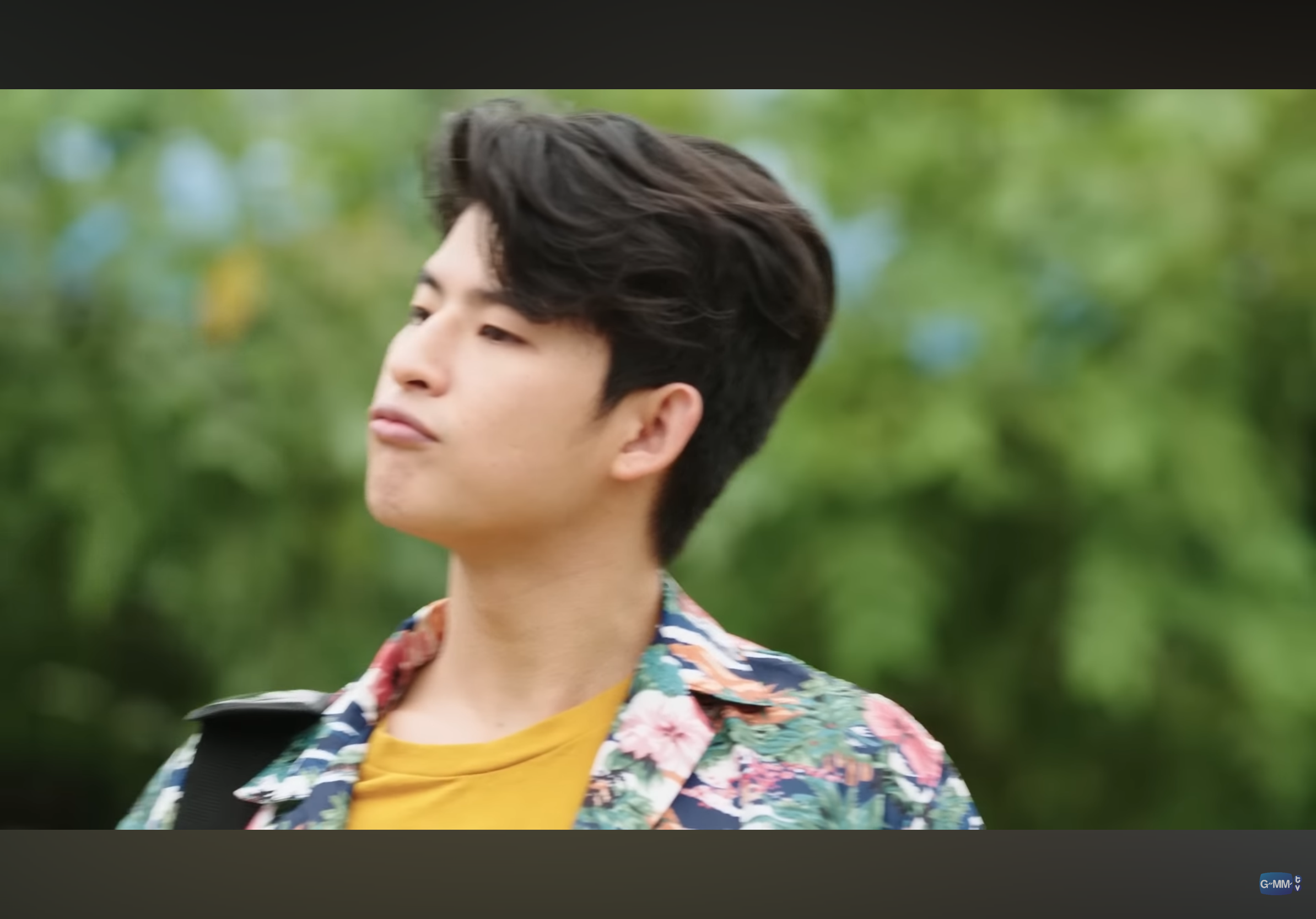 a screenshot of a young, asian man with dark hair styled to the side. he is wearing a floral shirt and has a confused expression on his face. the background is a blurry, green, outdoor setting.