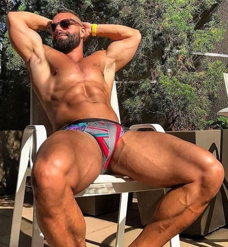 a muscular, tanned man with a beard and sunglasses is sitting on a white chair outdoors, wearing colorful patterned swim briefs. he has his arms behind his head, relaxed and enjoying the sun.  the background suggests a sunny vacation setting with lush green vegetation. his physique is very welldefined and muscular.
