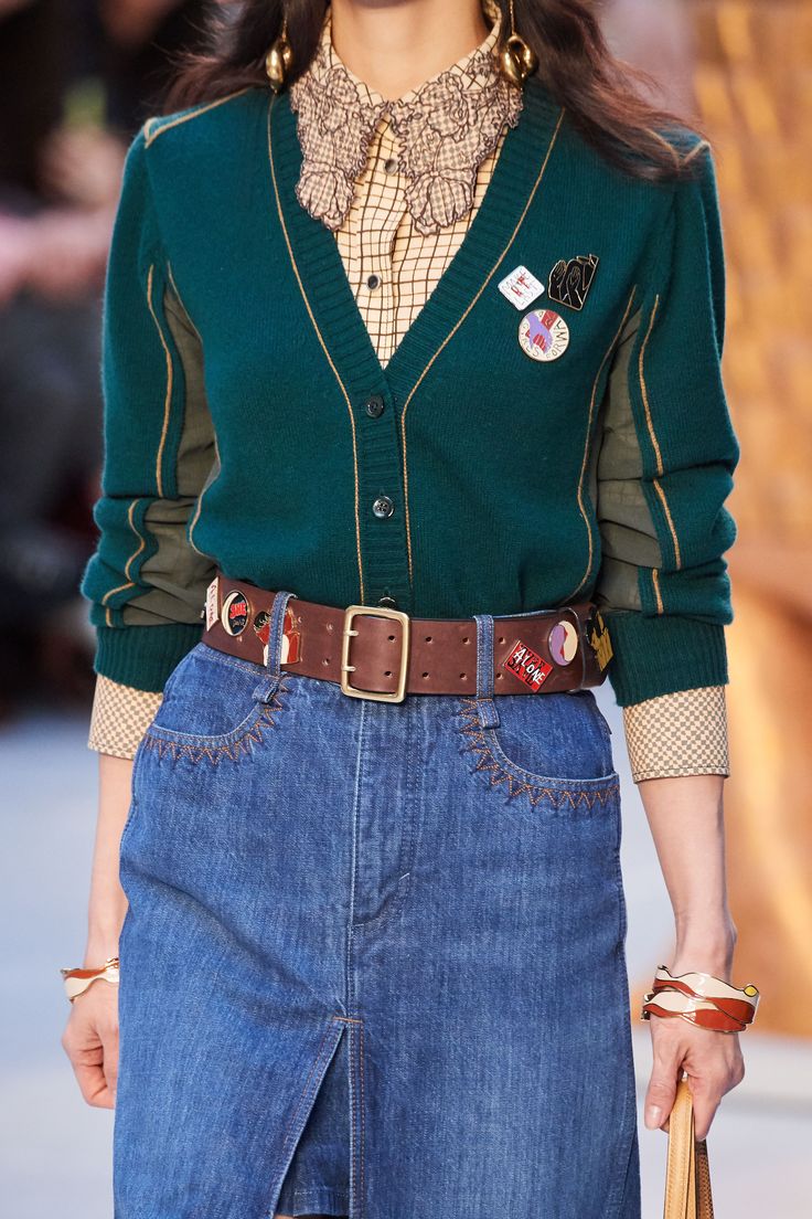 loseup shot of a model on the chloe fall/winter 2023 runway. she's wearing a teal green cardigan with gold detailing, a denim skirt with decorative stitching, a brown leather belt adorned with pins and badges, gold earrings, and bracelets. the overall style is a mix of vintage and contemporary elements.  a portion of a handbag is visible in her hand.