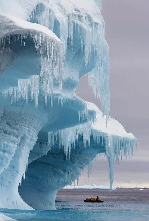 a massive iceberg covered in long icicles stands in the frigid waters of the arctic or antarctic. a small boat carrying people is visible near the base of the iceberg, providing scale to its immense size. the sky is overcast, creating a cold and serene atmosphere. the scene depicts the beauty and fragility of the frozen landscape.
