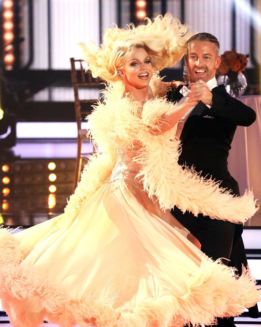 a woman in a glamorous peachcolored gown with large feather embellishments dances with a man in a classic black suit on a brightly lit stage.  her blonde hair is styled in a dramatic updo, and they are both smiling, suggesting a joyous and entertaining performance. the stage features a hint of art deco design elements in the background.