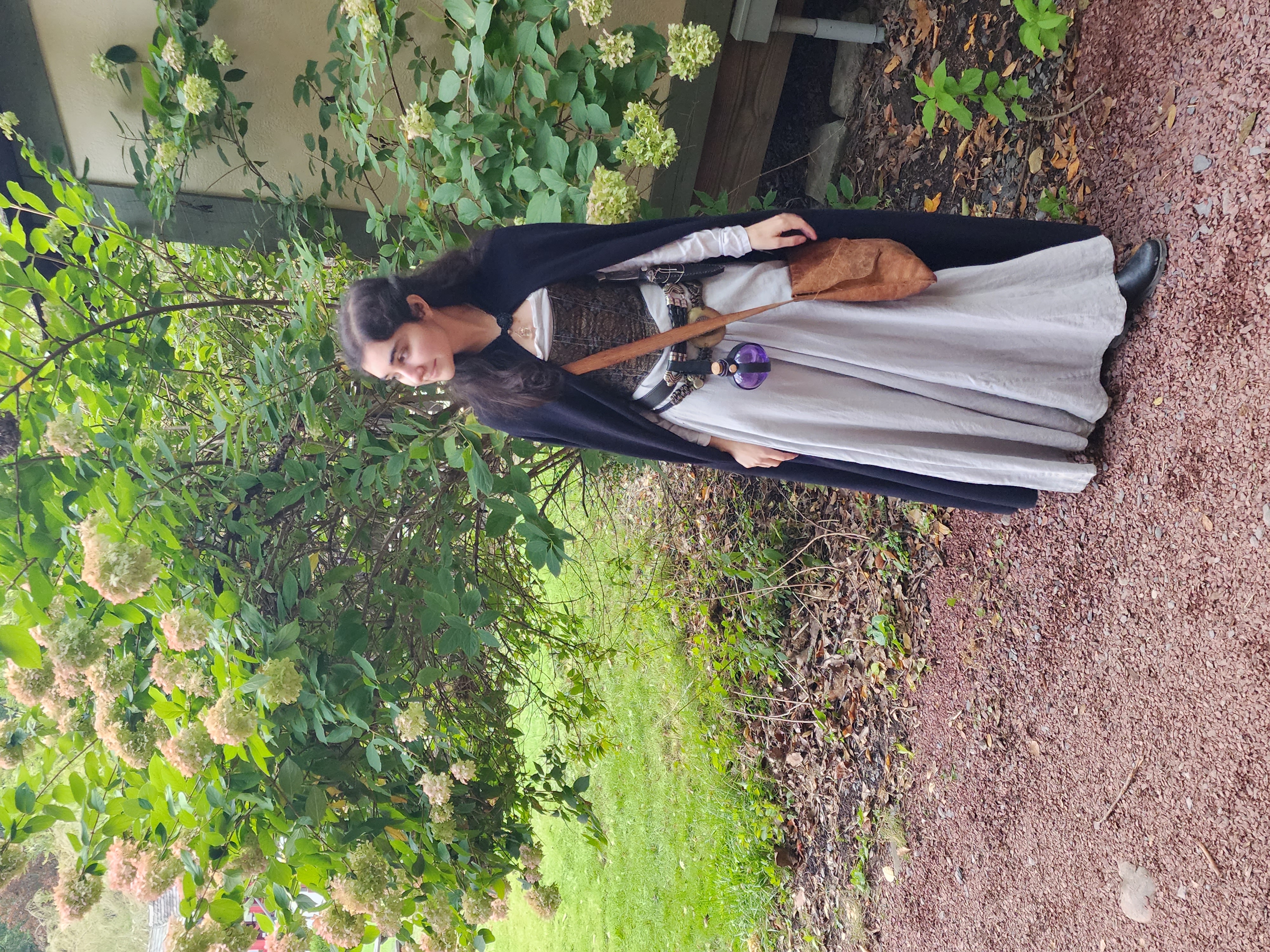 a young woman in a medievalstyle costume stands outdoors in a garden. she is wearing a long, light gray skirt, a brown corset, and a black cloak. she also has on a brown belt with a small, round, purple pouch hanging from it, and carries a small brown satchel.  she is smiling and looking away from the camera.  the background features a lush green shrub with many hydrangea blooms. the ground is covered with small, reddishbrown stones.