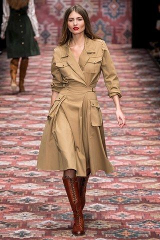 a fashion model walks down a runway wearing a tan, militarystyle midi dress with pockets.  the dress is belted at the waist and has a wrapstyle front. she's wearing long, brown, laceup boots. the runway has a patterned carpet in muted autumnal colors. another woman in a dark green dress is visible in the background, slightly out of focus. the overall aesthetic is vintage or retroinspired, with an autumnal feel.