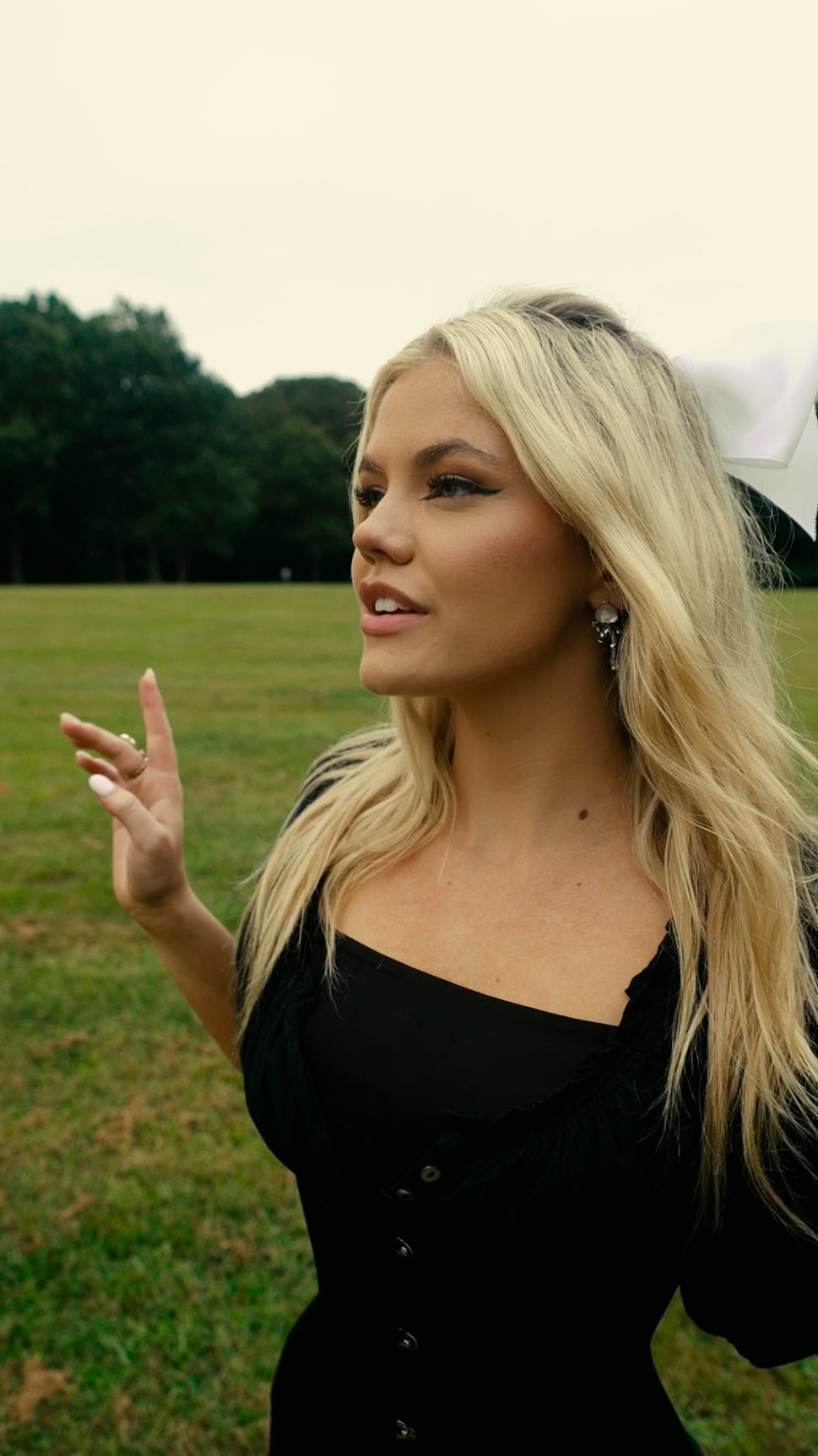 a woman with blonde hair stands in a meadow, wearing a black dress with buttons and a white bow in her hair. she is wearing jewelry, including a ring and earrings. in the background, there are trees and grass.