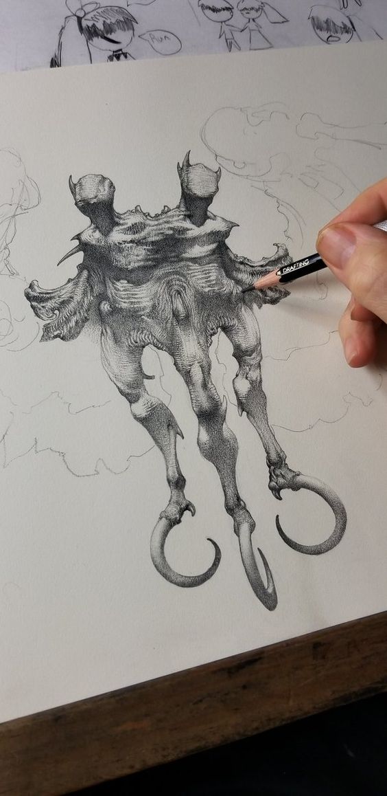 the image shows an inprogress pencil drawing of a twoheaded monstrous creature with wings and clawed feet. the creature appears to be floating. the drawing is being done on a large sheet of white paper on a wooden table. the artist's hand is visible in the image holding a pencil and actively shading a section of the creature's wing. other sketches of smaller, simpler figures are visible in the background at the top of the page. the lighting in the image is bright and diffused, suggesting natural light coming from a window.
