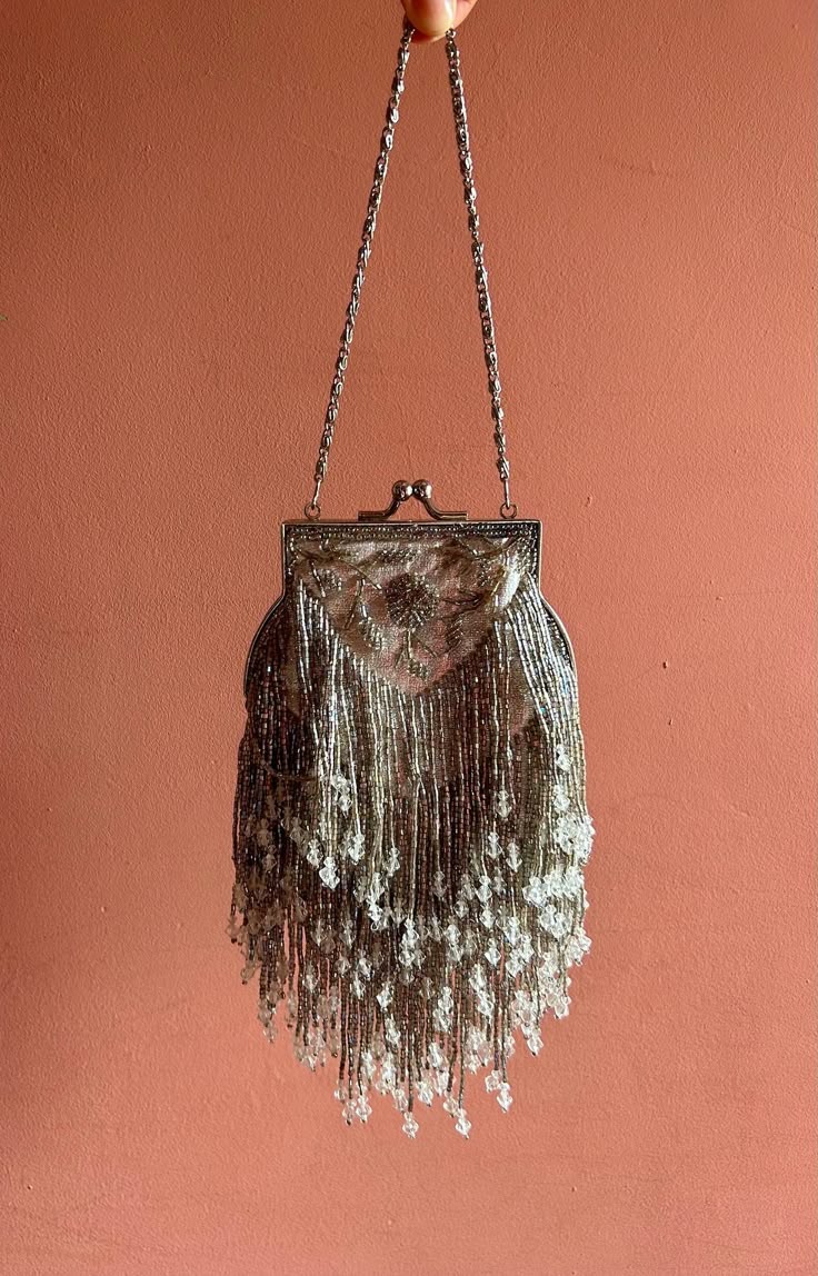 a silver beaded flapper bag is hanging against a peachcolored wall. the bag has a beaded handle and a silver frame. the body of the bag is covered in silver beads, and long silver beaded fringe.