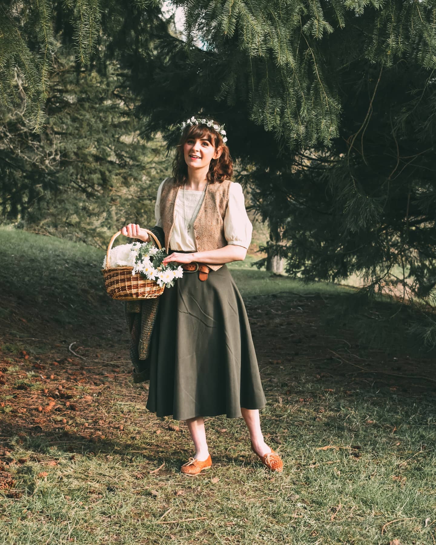 a young woman in a vintagestyle outfit stands in a forest, smiling at the camera. she is wearing a green skirt, a brown vest, and a white blouse. she carries a basket of flowers. she wears a floral headband and brown shoes. the scene is peaceful and idyllic.