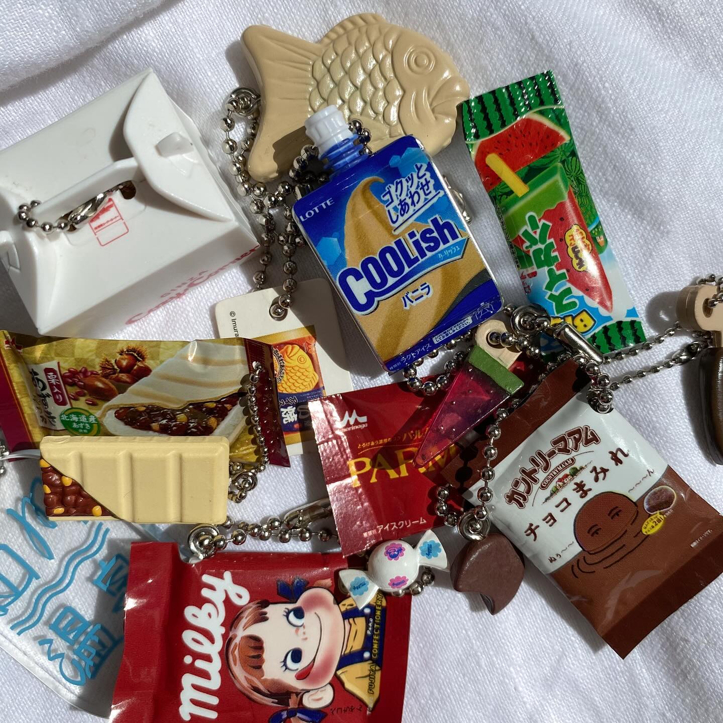 a closeup image shows several miniature foodthemed keychains spread across a white cloth. these include replicas of popular japanese snacks and treats, such as a "milky" candy package, a "coolish" drink pouch, a "taiyaki" fishshaped pastry, a "parm" ice cream bar wrapper, and a "country ma'am" chocolate cookie bag. each item is attached to a silver chain. the image has a slightly overexposed look, with the white cloth appearing bright.
