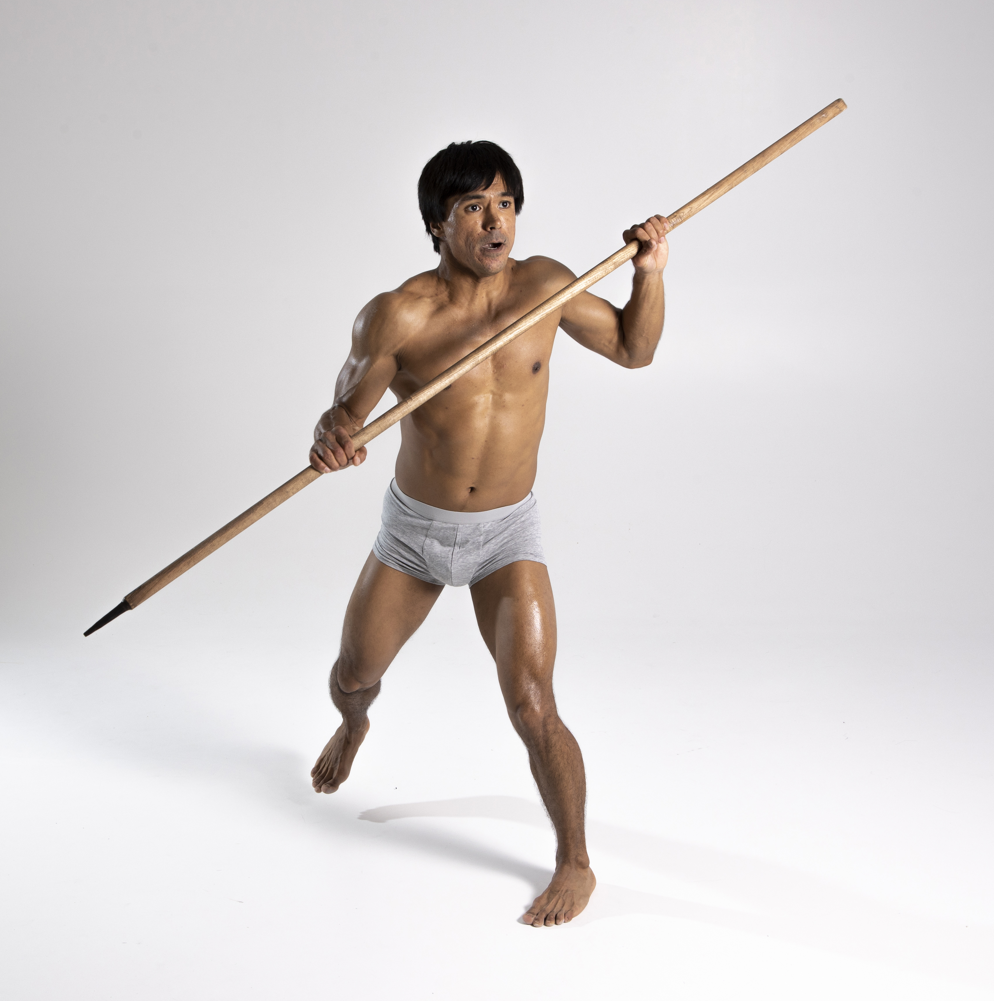 a muscular, shirtless man is running barefoot across a white studio background. he is wearing gray underwear and holding a long, wooden spear horizontally in front of him. the spear has a pointed tip. the man is looking forward with an intense expression.