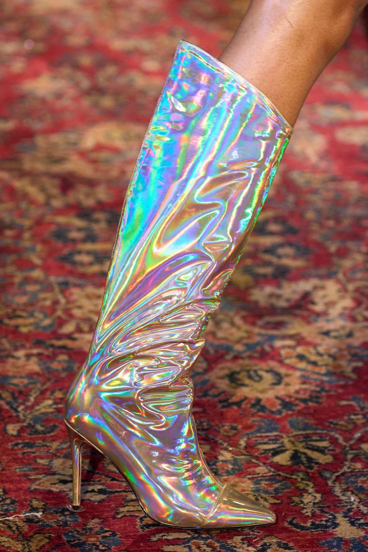 loseup view of a woman's leg wearing a pair of iridescent, kneehigh boots with stiletto heels. the boots have a holographic, rainbowlike sheen and a slightly wrinkled texture. the boots are shown against a richly patterned carpet with deep reds, golds and other rich tones.  the overall style is glamorous and eyecatching.