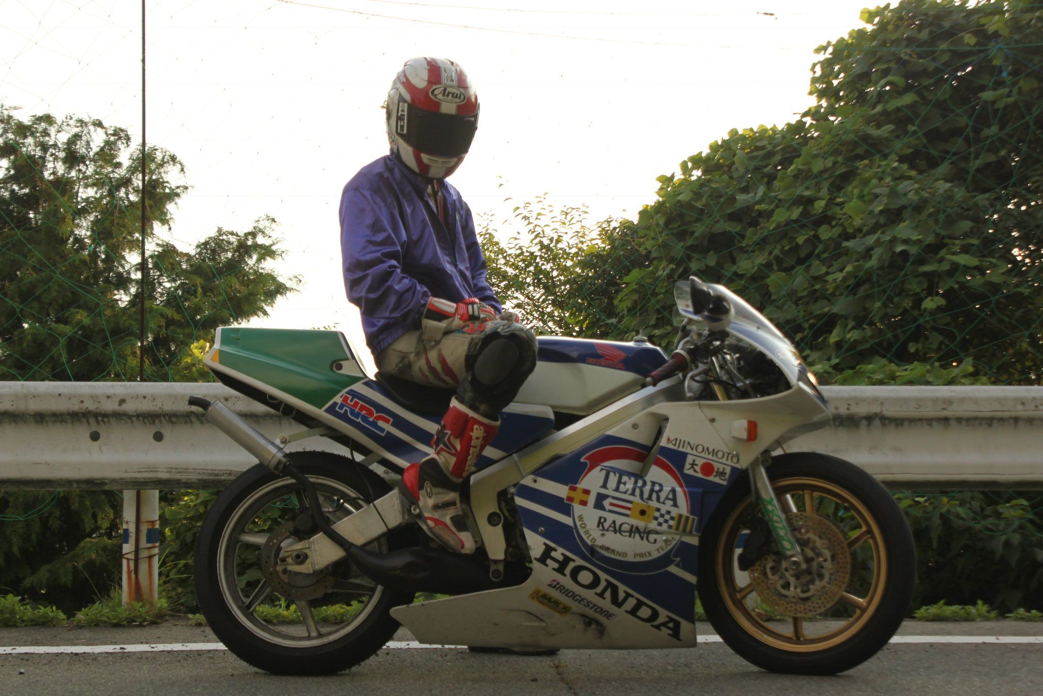 a motorcycle racer in full leathers is stopped on a road while wearing a red, white, and blue helmet. the bike is a white, blue, and red honda with the words "terra racing" and "bridgestone" printed on the side. the bike is parked in front of a guardrail, the racer is wearing a blue jacket, and the background is a forest with a road winding through it.