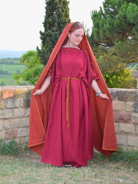 the image shows a woman wearing a historically accurate recreation of a roman stola (a long, simple dress) and palla (a shawl or cloak). the stola is a deep crimson red, and the palla is a burnt orangered.  the woman is wearing a simple headband. she is standing outdoors, with a stone wall in the background. a picturesque landscape is visible behind the wall, suggesting a historical or reenactment setting. the overall look is elegant and simple, representing ancient roman attire with care for detail in fabric and design.