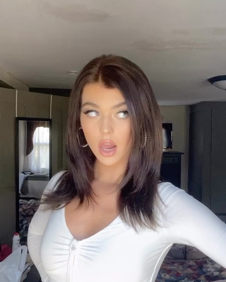 a portrait of a woman with brown hair, wearing a white top and earrings, looking away with her mouth slightly open. the image is taken indoors, possibly in a bedroom, with a mirror visible in the background.