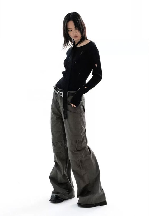a full shot of a young woman model posing against a white background. she is wearing a black distressed longsleeve sweater and dark grey wideleg cargo pants. her hair is dark and styled straight. she is wearing black shoes. the overall style is casual and contemporary. the image is welllit and shows the details of the clothing.