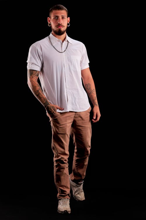 a fullbody studio shot of a man with tattoos, wearing a white shirt, brown pants, sneakers, and a silver necklace, standing against a black background. he has short hair, a beard, a mustache, and earrings. he has one hand in his pocket.