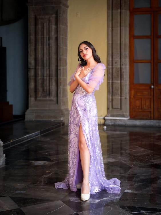 a woman wearing a purple sequined gown with a thighhigh split, stands inside a building with stone columns and a marble floor. she has her hands crossed in front of her chest and is looking at the camera.