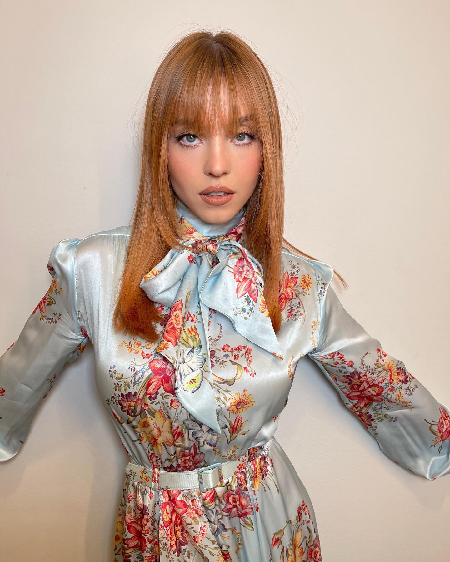closeup shot of a young woman with long, straight ginger hair and bangs, wearing a light blue floral satin dress with a bow tie collar. the dress has a long sleeve and a belted waist. she is looking directly at the camera.  the background is a plain, light beige/offwhite wall. the overall impression is one of elegance and sophistication.