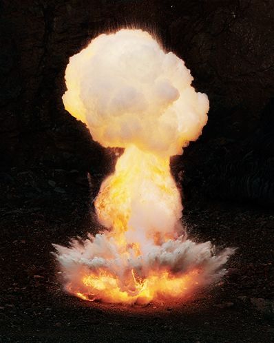 the image shows a large explosion with a mushroom cloud rising against a dark, possibly rocky, background. there are flames and smoke visible near the ground, indicating the point of detonation. the explosion conveys a sense of power, danger, and destruction.