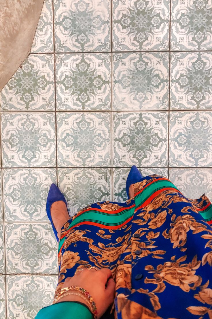 a highangle, closeup shot captures a woman's feet clad in blue suede pointedtoe heels. she's wearing a vibrant blue dress adorned with a gold floral pattern, accented by a teal green, red, and orange striped band at the waist.  her wrists are adorned with gold bracelets. the woman is standing on a beautifully patterned floor composed of square tiles in shades of offwhite and tealgreen, featuring an intricate, repeating design. the image's focus is on the interplay of colors and textures between the clothing and the floor.