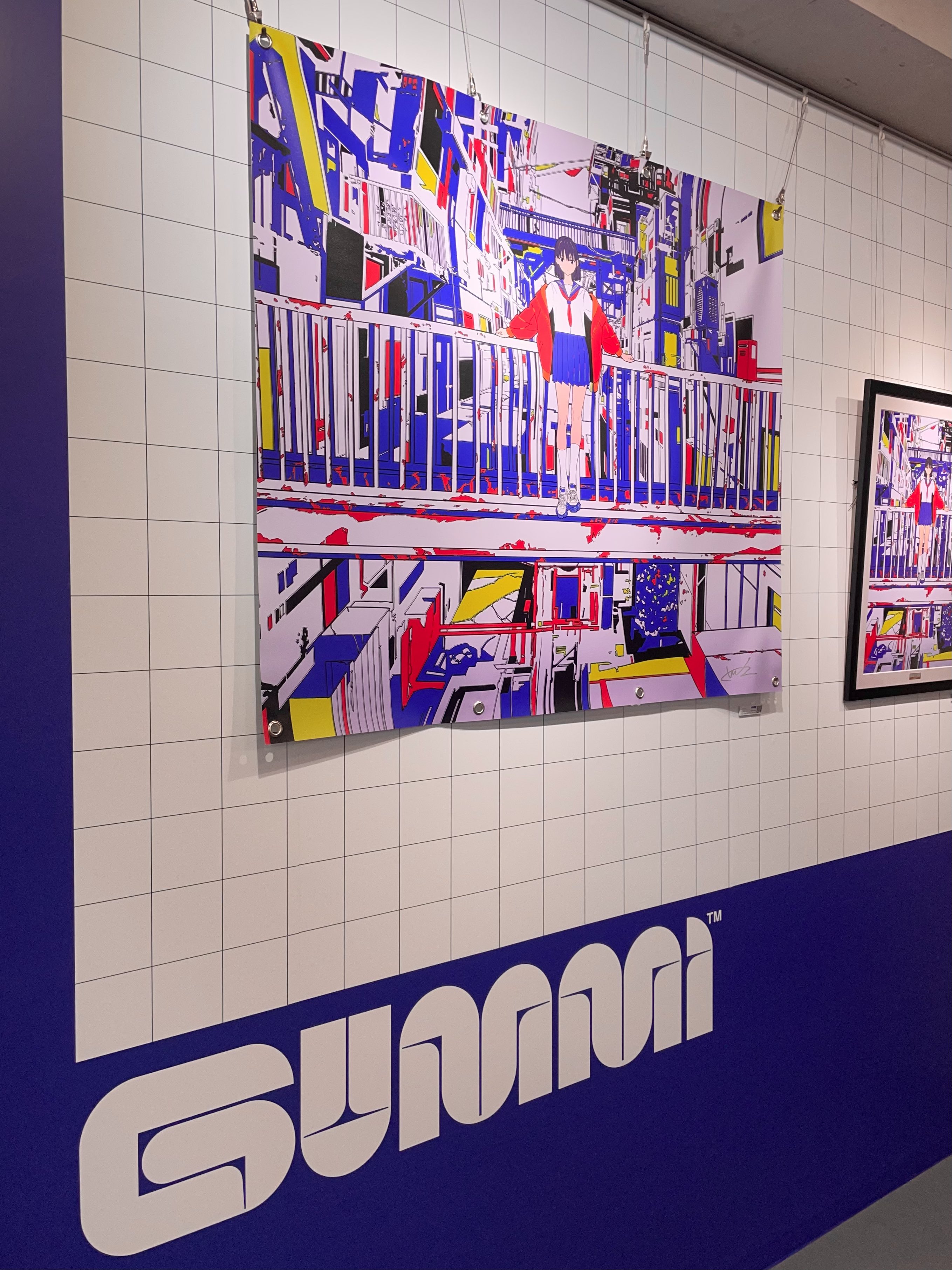 this image showcases a vibrant, graphic artwork displayed in a gallery setting. the piece, featuring bold colors and geometric shapes, depicts a girl standing amidst a cityscape. the artwork hangs on a white tiled wall with the name "sunnei" prominently displayed in white and light blue on a darker blue section of the wall. a second, smaller artwork is partially visible on the right. the gallery is welllit with artificial lighting.