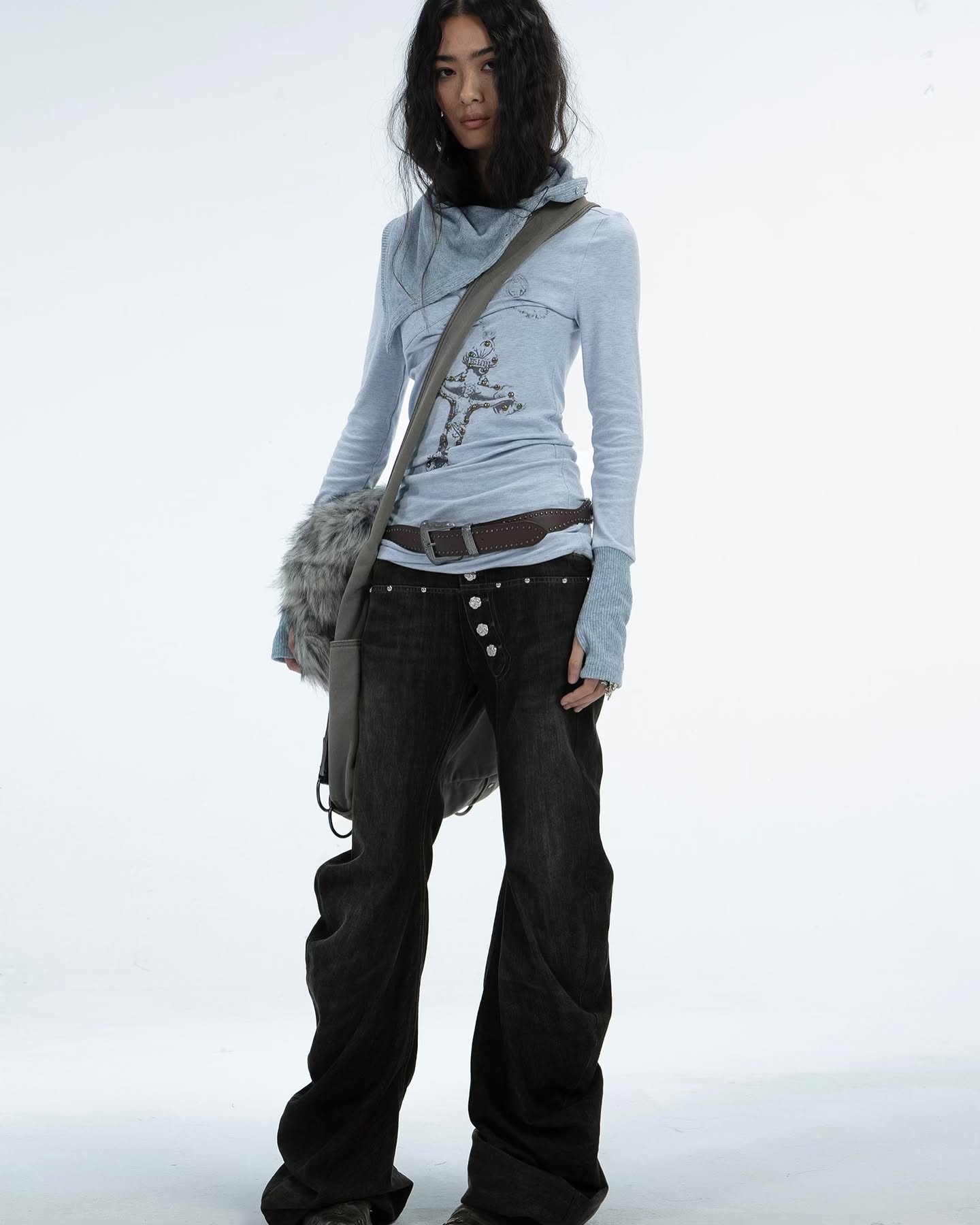 a full shot of a young woman model posing against a white background. she is wearing a light blue longsleeved shirt with a cowl neck and a graphic print. the shirt is tucked into darkgrey wideleg jeans. she is also wearing a grey furry shoulder bag and a dark grey belt with silver buttons. her dark hair is slightly tousled, and she has a neutral expression. the overall style is casual and relaxed.