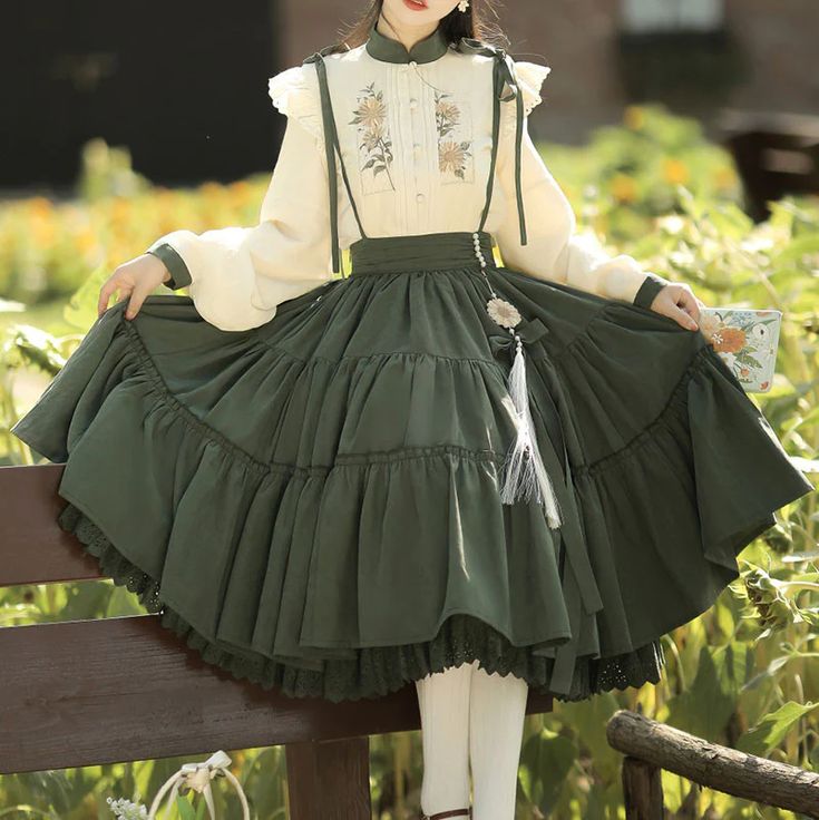 a woman wearing a longsleeved, creamcolored blouse with floral embroidery paired with a long, flowing dark green skirt. the skirt is tiered with ruffles at the hem and features a decorative tassel. she is posing outdoors in a garden setting and is holding the edge of her skirt, showcasing its volume. she is also carrying a small bag. the overall aesthetic is romantic, feminine, and evocative of vintage fashion and natureinspired styles.