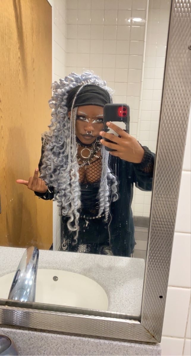 a woman with silver hair is standing in a bathroom taking a selfie. she is wearing black clothing, a nose ring and silver eyeliner.  a white porcelain sink with a silver faucet is in front of her.  she is looking directly at the camera.