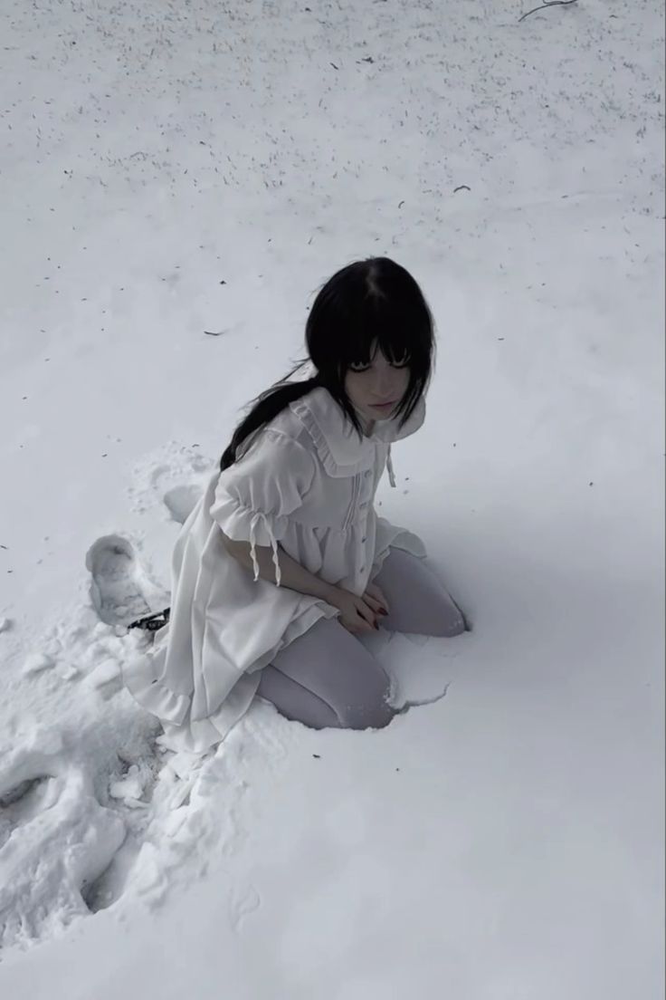 a young woman with long black hair, pale skin, and dark eyes is kneeling in the snow. she is wearing a white dress with ruffled details. the setting is a winter scene with a blanket of snow covering the ground. footprints are visible in the snow around her. the overall mood is ethereal and serene.