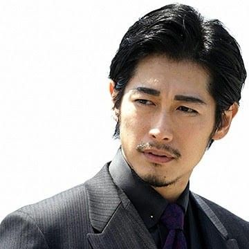 the image is a headshot of a japanese man with short black hair, a goatee, wearing a dark gray suit and a purple tie. he is looking down and to his right. the background is white and the lighting appears to be studio lighting.