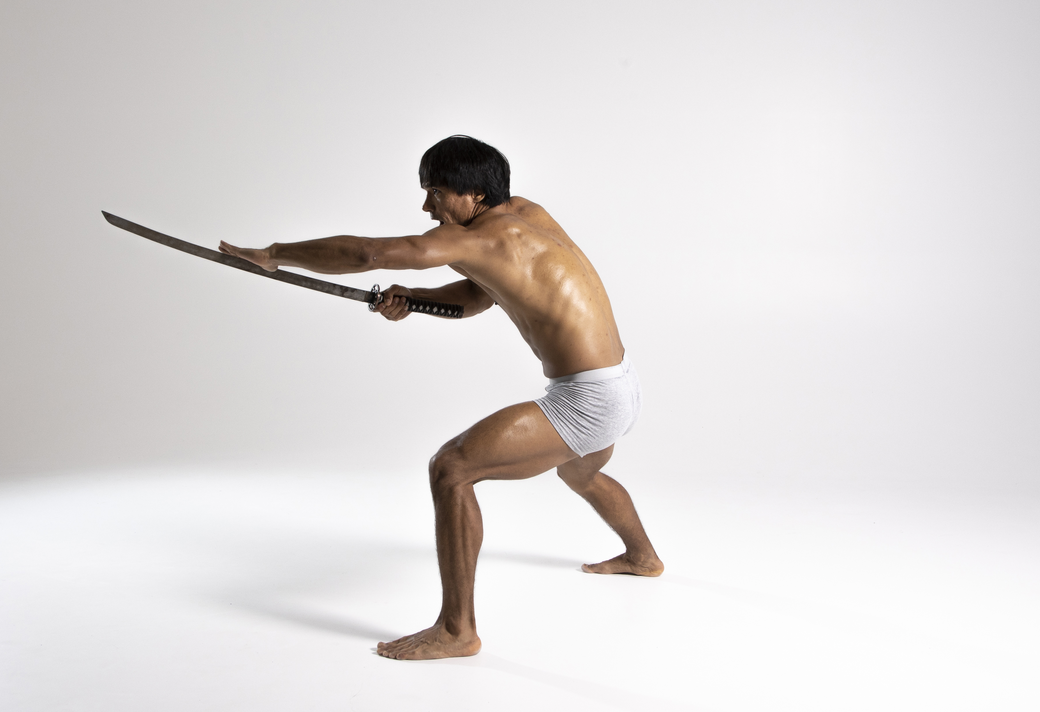 a barechested, athletic man is holding a katana in a fighting stance against a white studio background.