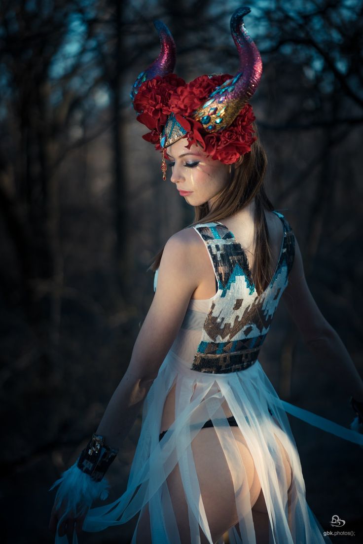 a young woman with elaborate makeup, horns, and a floral headdress stands in a dimly lit forest. her costume is a flowing white garment with a decorative bodice. she embodies a mystical or fantasy character, and the overall mood is ethereal and slightly melancholic. the image is taken from slightly behind her, capturing her gaze directed downwards.