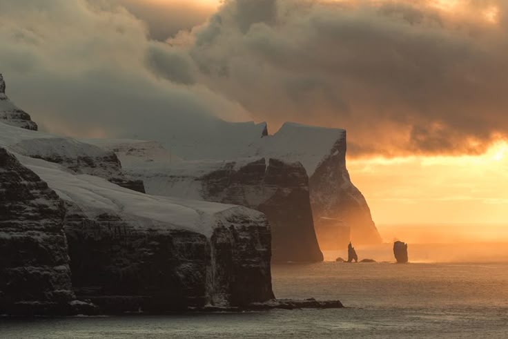 the image shows a landscape featuring snowcovered mountains and cliffs near the sea, with a sunset and clouds in the background. the scene is likely in the faroe islands, possibly vágar, and includes rock formations like drangarnir and tindhólmur.