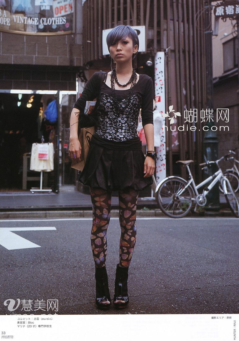 a young woman with a stylish, edgy look poses on a city street. she wears a black corset dress, butterflypatterned tights, and platform boots. her short hair is a striking shade of blue. the background features a vintage clothing store and parked bicycles, creating a casual, urban atmosphere.