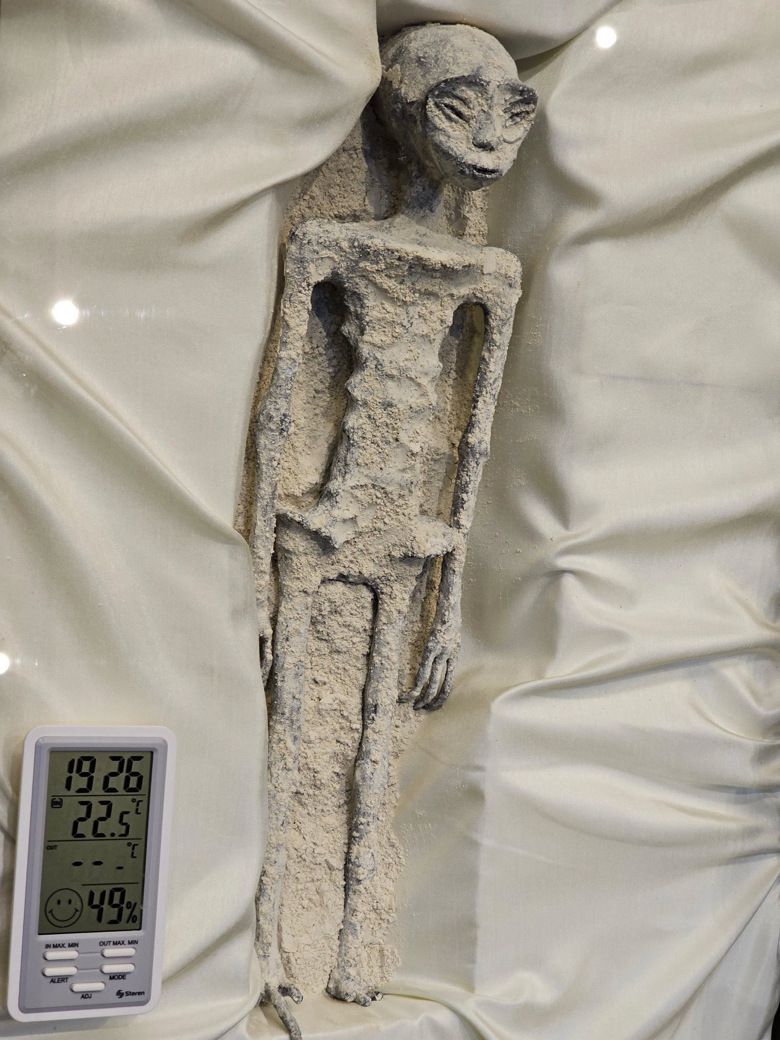 the image shows a mummified human body lying on a white cloth, encased in what appears to be a layer of sand or other preservative material. the body is positioned with its arms at its sides and its head turned slightly to the right. the skeletal structure of the mummy is clearly visible, indicating a significant degree of preservation. a digital thermometer is placed to the left of the mummy's feet, monitoring the temperature and humidity levels for its preservation. the setting suggests that the mummy is part of a museum exhibit, as indicated by the controlled environment and careful presentation.