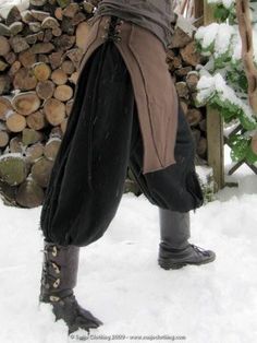 this is a picture of a person wearing a medieval inspired outfit in the snow. they are wearing brown leather boots, a dark brown tunic with a brown leather belt and brown pants. they are standing in the snow, there are trees and branches behind them. the picture has a rustic feel.