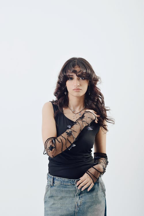 a teenage woman is posing for a studio shot. she is wearing a black top, low rise jeans, and black butterfly gloves. she also has on a couple of necklaces. she has long wavy hair and bangs.