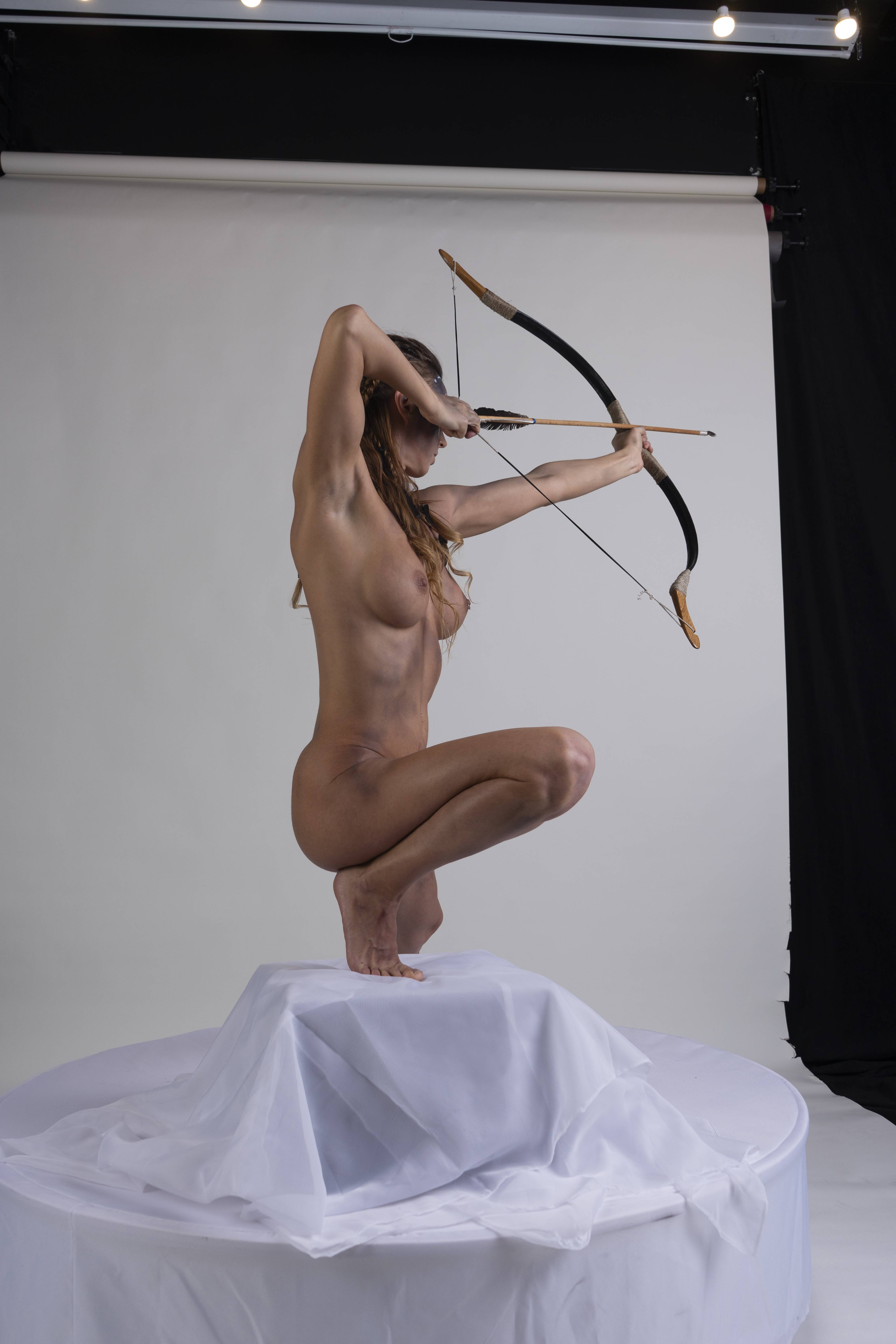a nude woman is crouching on a white draped platform, holding a bow and arrow, as if she is about to shoot. the background is a white backdrop with lighting equipment visible.