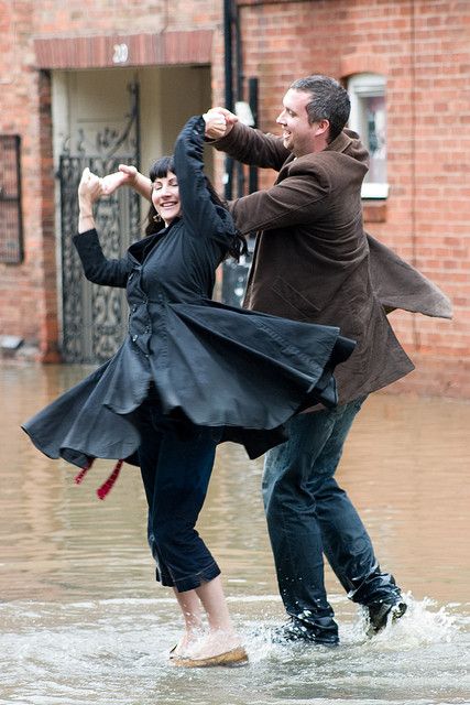 a couple dances in a flooded street on a rainy day. the woman is twirling in a black coat and jeans, while the man, wearing jeans and a brown jacket, is holding her hand and smiling. the street is flooded with water, creating a playful and romantic atmosphere. they are both happy and seem to be enjoying the rain and the opportunity to dance together.