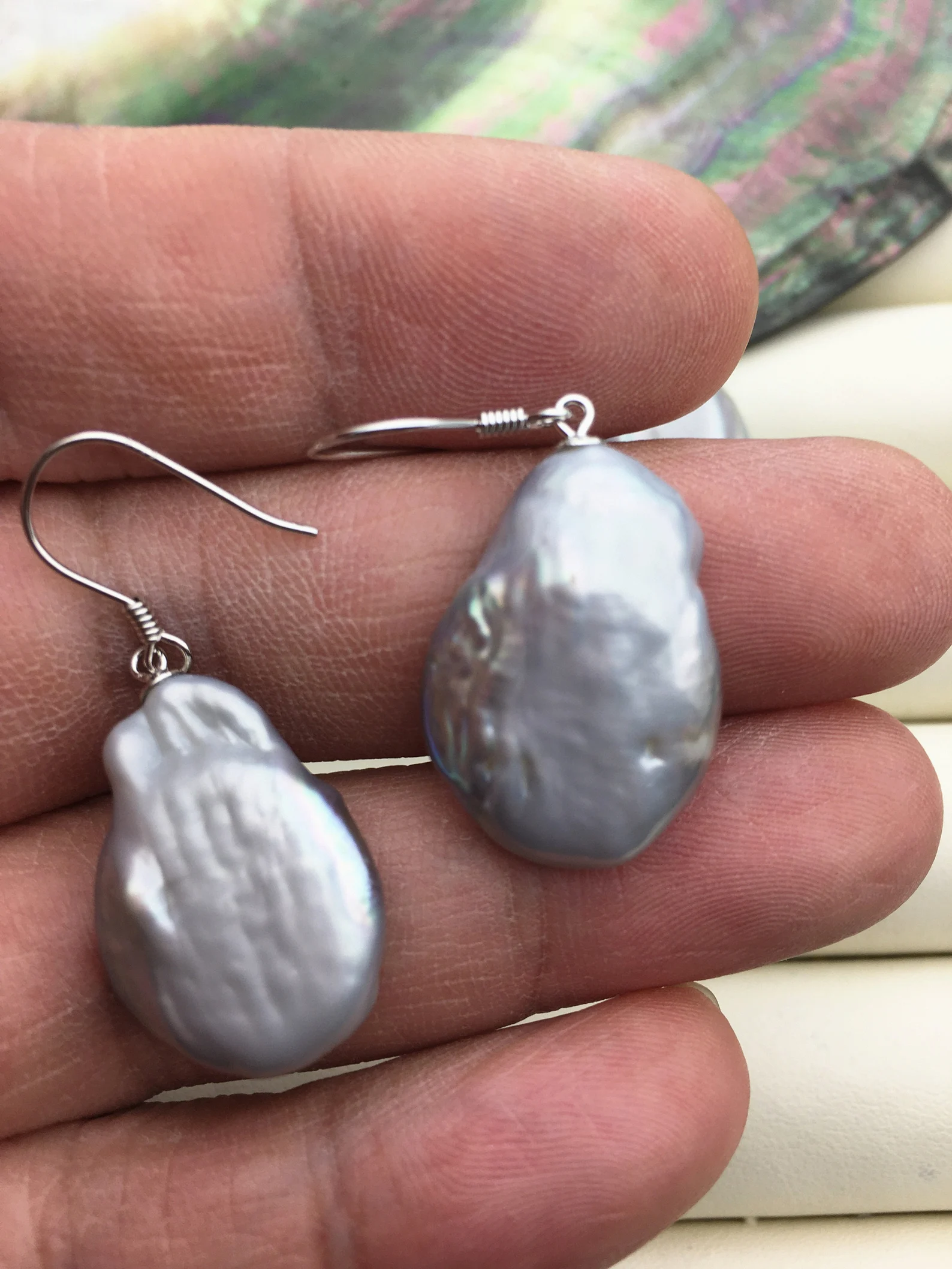 a person is holding a pair of gray baroque pearl earrings in their palm. the earrings have a simple silver hook and are set with large, irregularly shaped pearls that reflect the light softly. the background features a blurred abalone shell, known for its iridescent colours and used in pearl cultivation. content: earringpeople: yesenvironment: indoorlighting: natural lightingperspective: close upkw: jewelry, pearl, baroque, earring, fashion, accessory, gemstone, elegant, style, design, silver, grey, natural, handmade, gift, luxury, beauty, abalone, shell, mother of pearl, iridescent