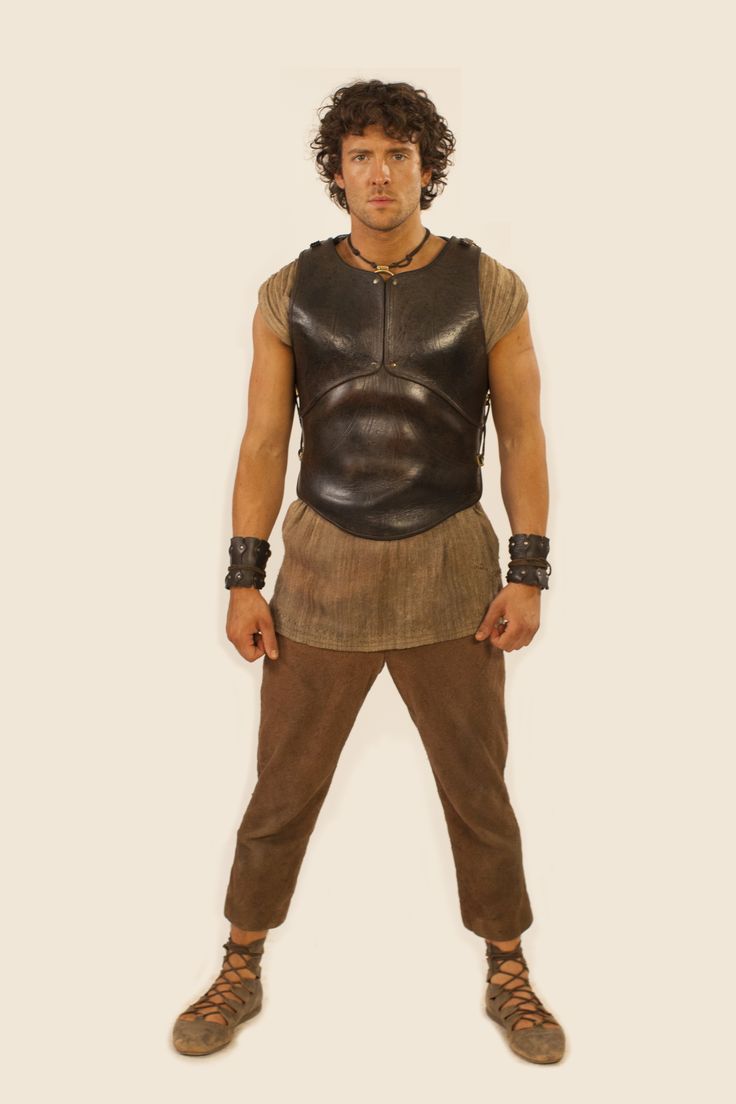 a full shot of a man standing, dressed in what appears to be ancient or medievalstyle clothing. he is wearing a brown leather cuirass over a tan tunic. his pants are brown, and he has brown leather sandals with laces. he is also wearing leather wristbands on both wrists and a simple necklace. the man has curly brown hair. he is standing against a plain light beige background.