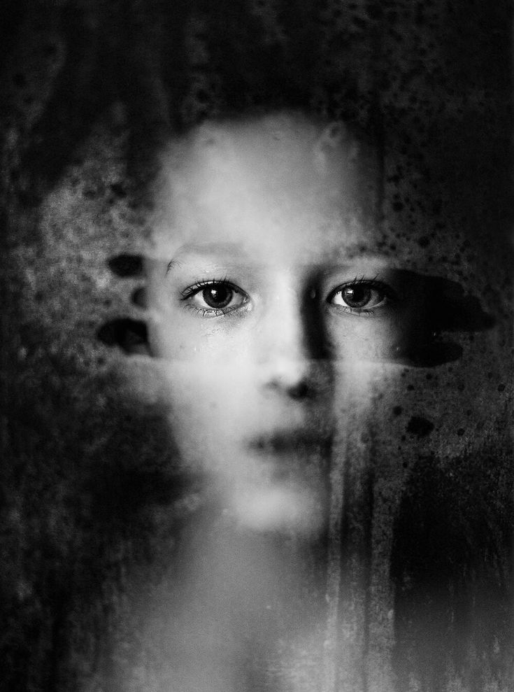 the image is a black and white closeup portrait of a young woman's face.  her face is partially obscured and blurred, as if seen through a dirty or foggedup window or glass. the background is dark and textured, adding to the mysterious and moody atmosphere.  her eyes are visible and seem to convey a sense of sadness or contemplation. the overall effect is artistic and evocative, leaving the viewer to interpret the emotion and story behind the image.