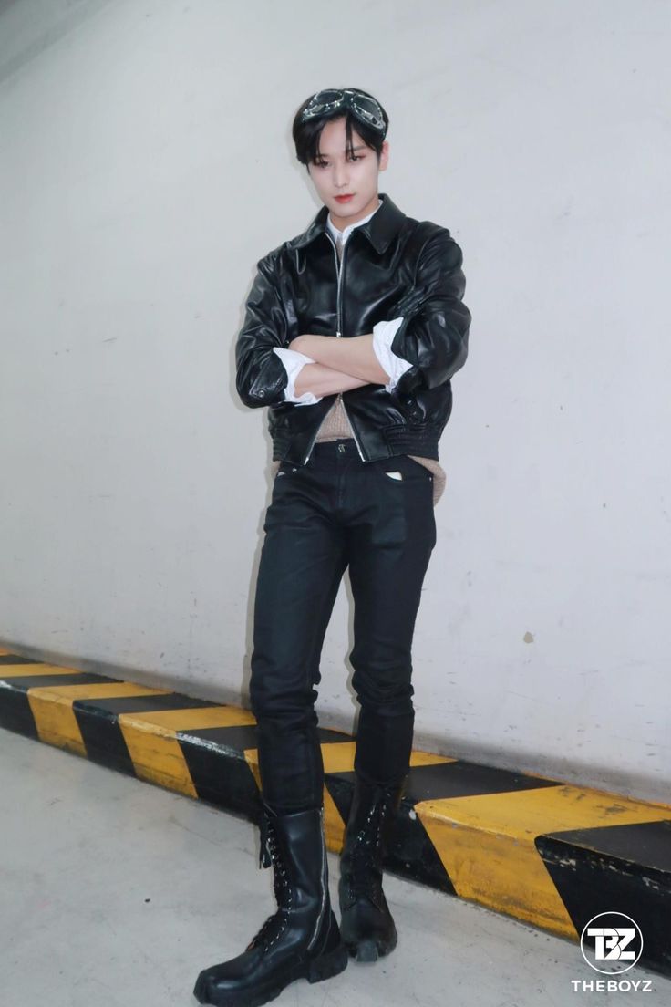 a fullbody shot of a man wearing a black leather jacket, black jeans, and black boots. he has his arms crossed and is posing in front of a white wall with a yellow and black hazard stripe along the bottom. he is also wearing a headband. the logo for the kpop group the boyz is in the bottom right corner.