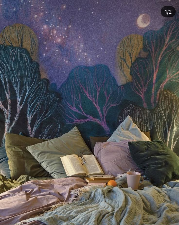 the image shows a cozy bed setup against a wall decorated with a drawing of a night sky, complete with stars, a moon, and trees. the bed is covered with pillows of various colors and a blanket. an open book with glasses on top rests on the pillows. a mug and an orange sit on the bed, suggesting a relaxing bedtime scene.