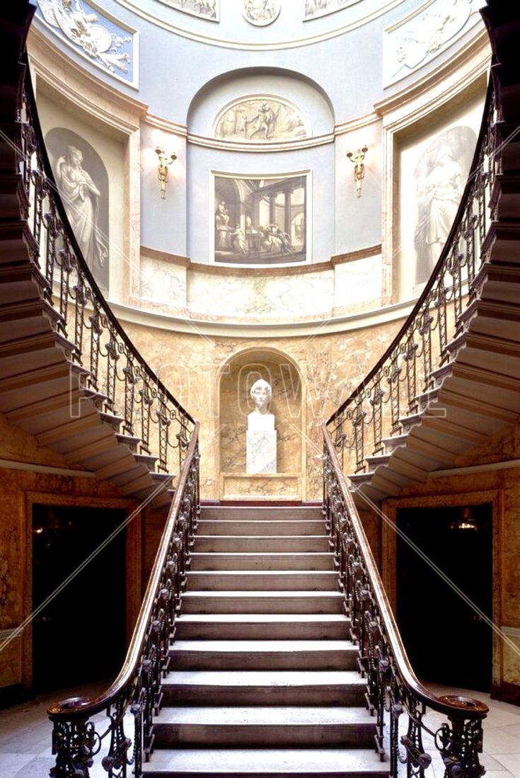 the image shows a grand, double spiral staircase in a luxurious, classicalstyle building. the staircase features an ornate wrought iron railing and winds around a central landing.  the walls are adorned with classicalstyle murals and paintings, while a marble bust sits centrally on the landing at the base of the stairs. the ceiling is a domed structure with more painted details.  the overall impression is one of opulence and historical significance.