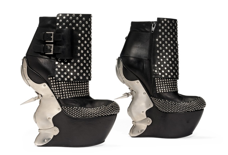 the image displays two black platform boots with a striking avantgarde design. the boots are made of black leather and are heavily adorned with silver studs. each boot features a high platform sole, a unique silver metal structure around the heel area, and various buckles and zippers. one boot has a buckled strap detail, while the other has a zipper on the side. they have a distinctive and edgy appearance.