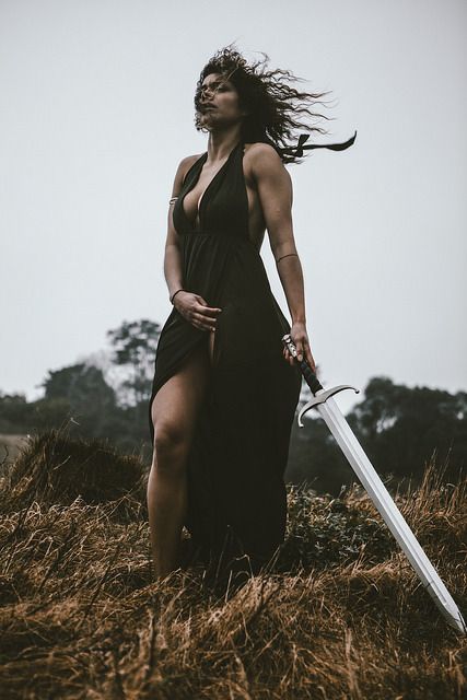 a woman stands in a field of grass, holding a sword.  her black dress blows in the wind, and she looks determined.  her hair is long and dark.  the photo is a beautiful and powerful image of a woman warrior.