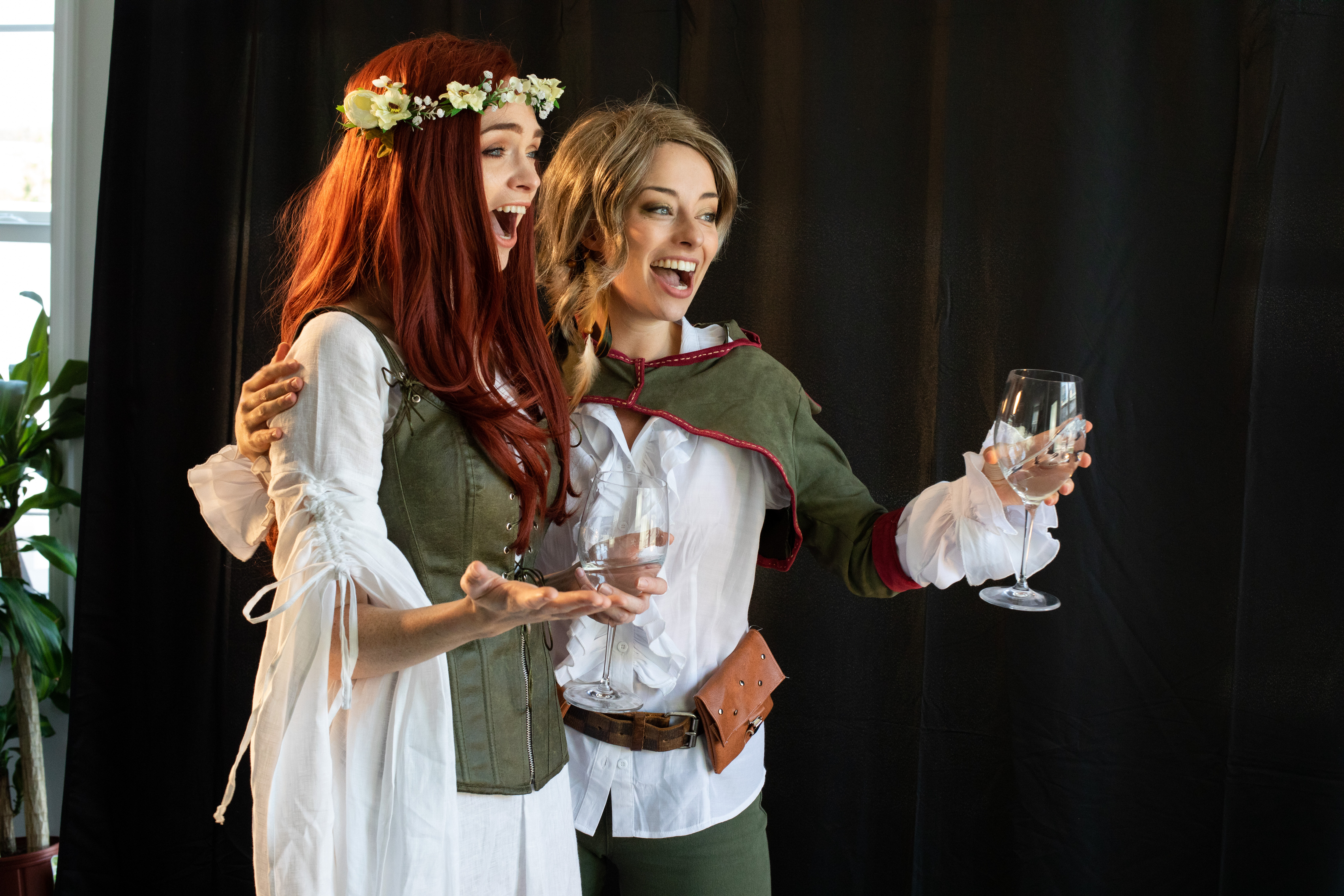two women dressed in fantasy cosplay laugh while holding glasses of white wine. the woman on the left has long red hair and wears a flower crown, a white dress and a green corset. the woman on the right has long blonde hair and wears a white shirt, a green vest and a brown belt. the background is black.